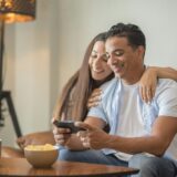 Happy young happy couple using smartphone social media apps at home, smiling husband and wife